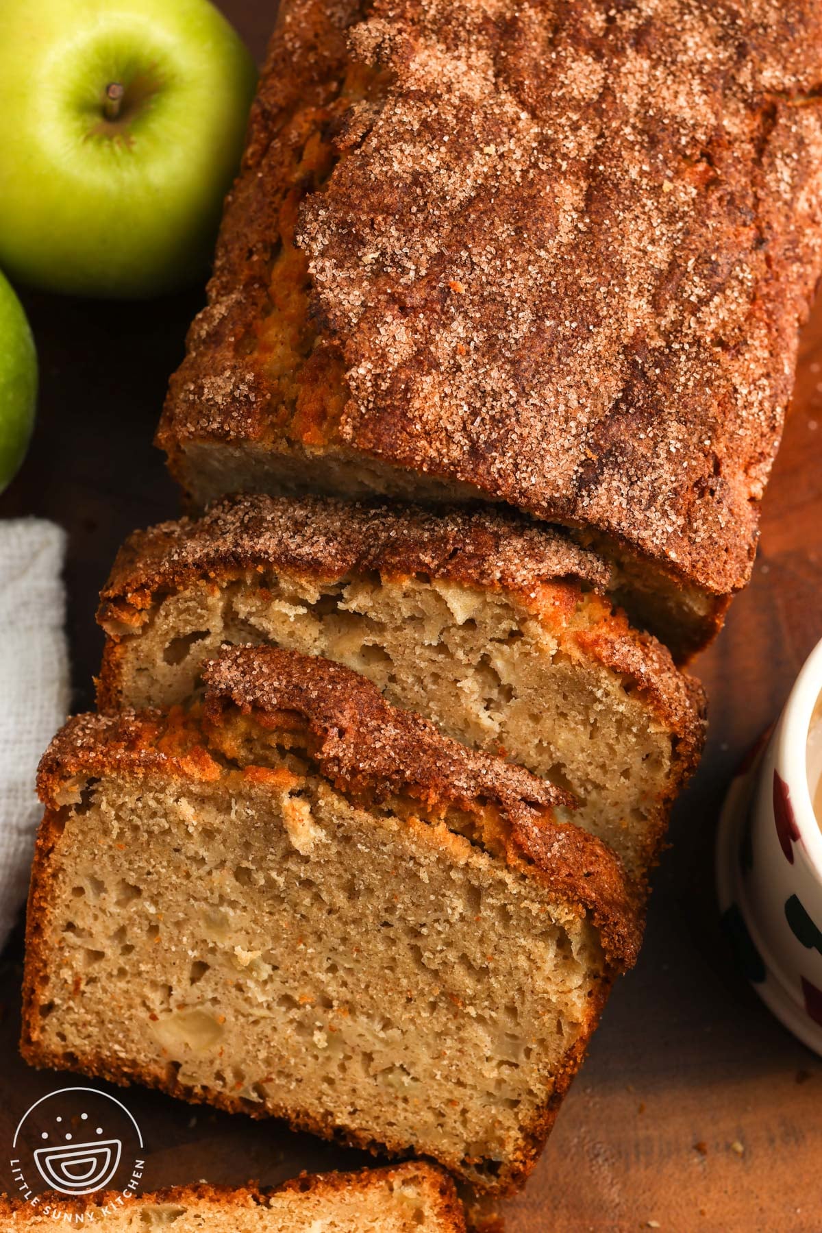 A sliced loaf of apple bread with apples in the crumb with granny smith apples on the left and a mug on the right.