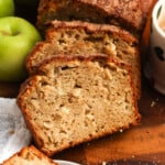 A sliced loaf of apple bread on a wooden cutting board. The slices show a tender crumb with apple pieces and a cinnamon sugar top. Next to the loaf are green apples and a mug of coffee.