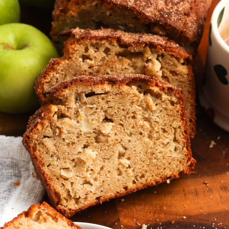 A sliced loaf of apple bread on a wooden cutting board. The slices show a tender crumb with apple pieces and a cinnamon sugar top. Next to the loaf are green apples and a mug of coffee.