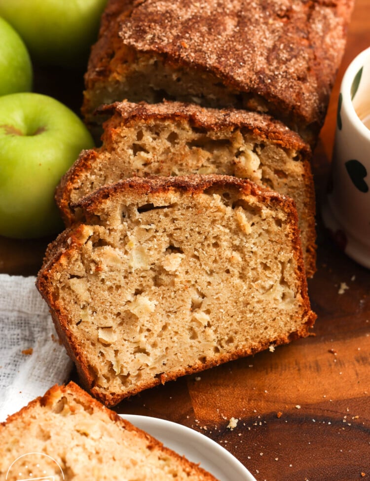 A sliced loaf of apple bread on a wooden cutting board. The slices show a tender crumb with apple pieces and a cinnamon sugar top. Next to the loaf are green apples and a mug of coffee.