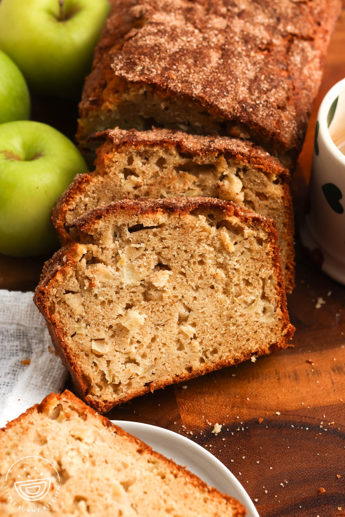 A sliced loaf of apple bread on a wooden cutting board. The slices show a tender crumb with apple pieces and a cinnamon sugar top. Next to the loaf are green apples and a mug of coffee.