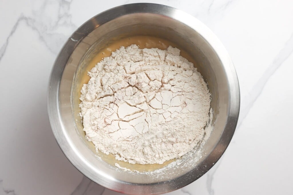 Dry flour added to wet ingredients in a metal bowl to make apple bread.