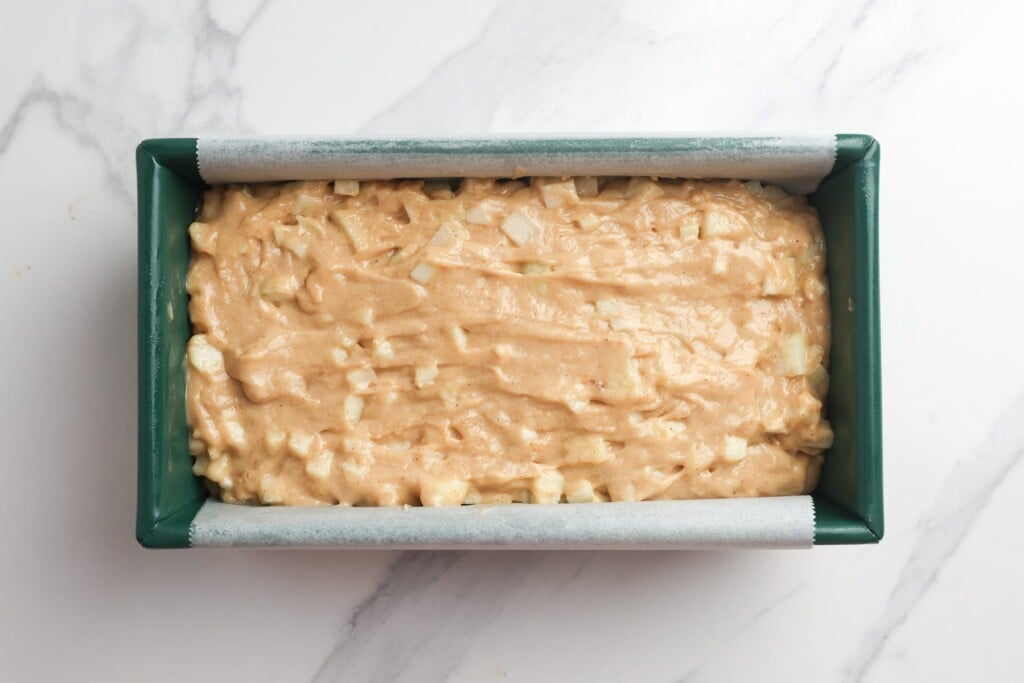 Apple bread batter with chunks of fresh apple in a green ceramic loaf pan that is lined on two long sides with parchment paper.