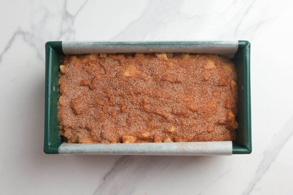 Cinnamon sugar topping spread over apple bread batter in a green loaf pan, before baking.