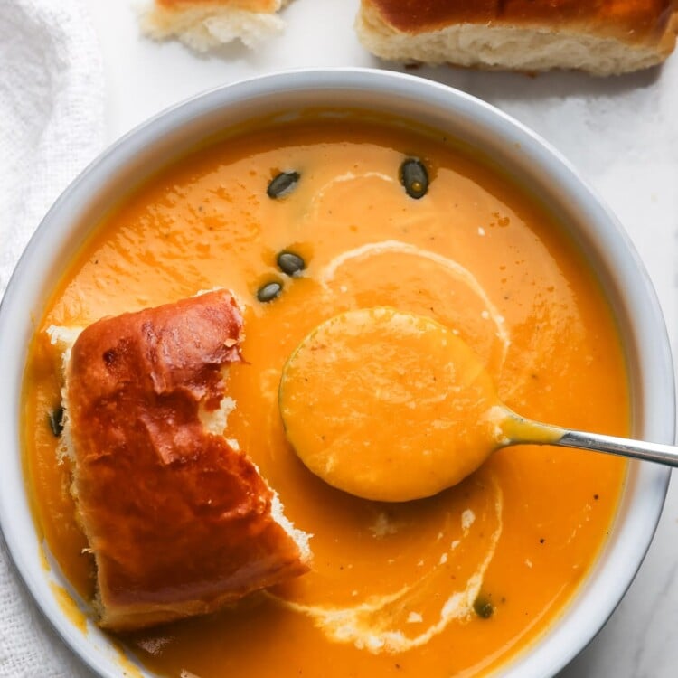 A white bowl of slow cooker butternut squash apple soup topped with a coconut milk swirl and pepitas, served with soft dinner rolls.