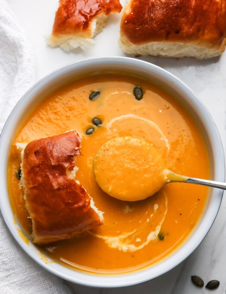 A white bowl of slow cooker butternut squash apple soup topped with a coconut milk swirl and pepitas, served with soft dinner rolls.
