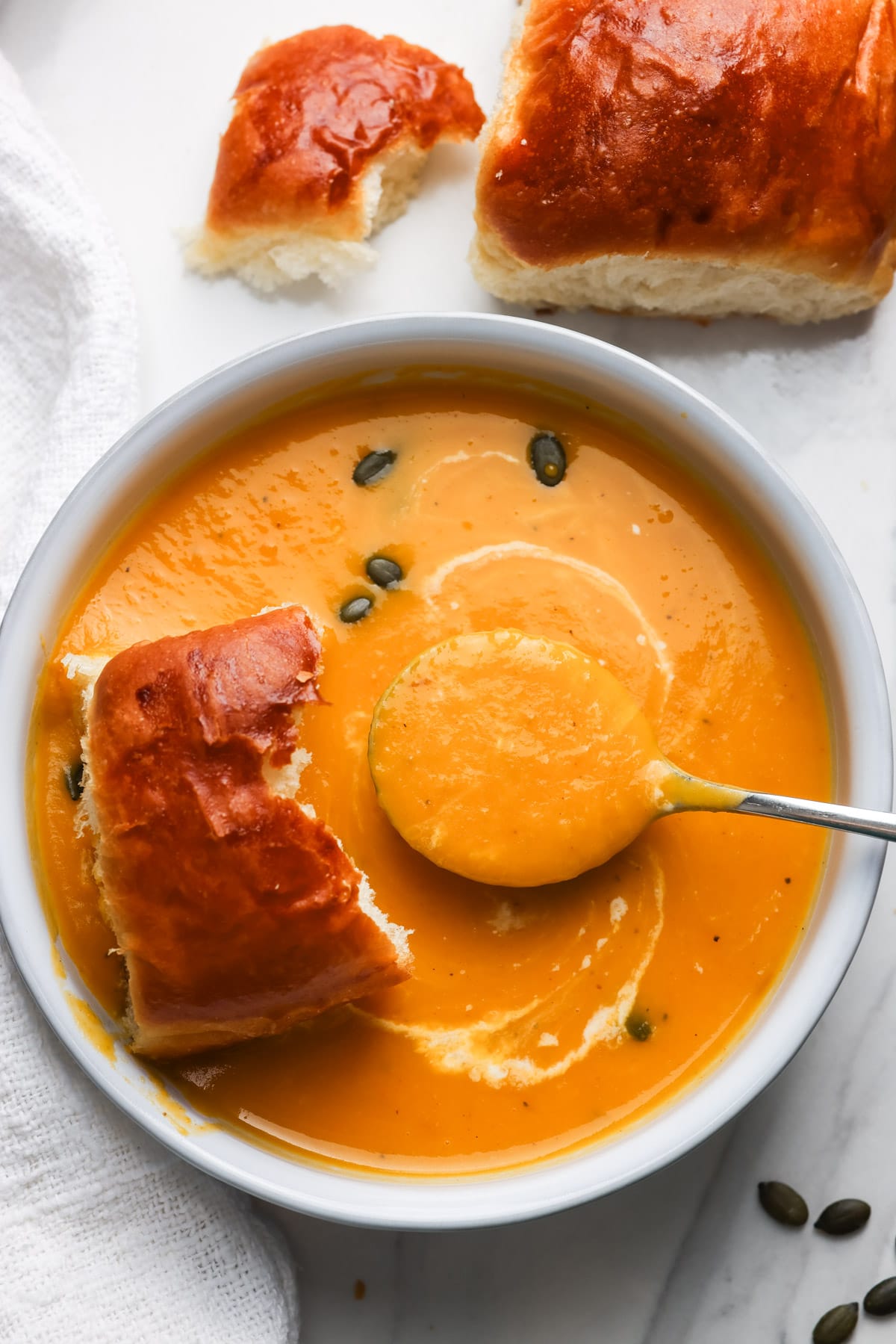 A white bowl of slow cooker butternut squash apple soup topped with a coconut milk swirl and pepitas, served with soft dinner rolls.