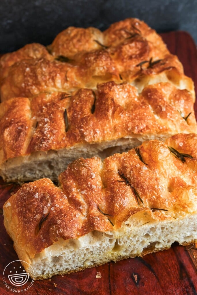 thin slabs of rosemary topped focaccia on a wood board, cut to show the open crumb inside.