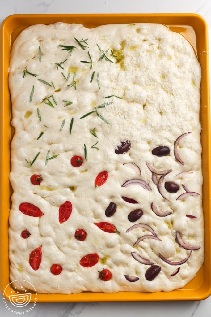 dimpled focaccia dough on a sheet pan before baking, topped in sections with rosemary, parmesan, clided cherry tomatoes, and olives and red onions.