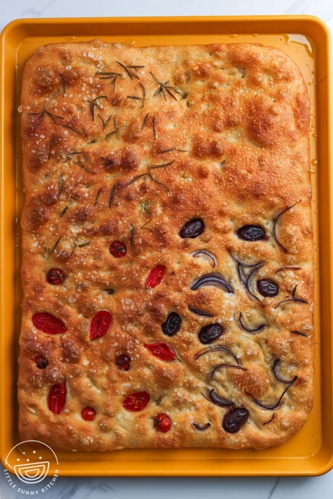 baked sheet pan focaccia with deep golden top showing four topping sections of rosemary with flaky salt, parmesan cheese, roasted cherry tomatoes, and olives with onion.