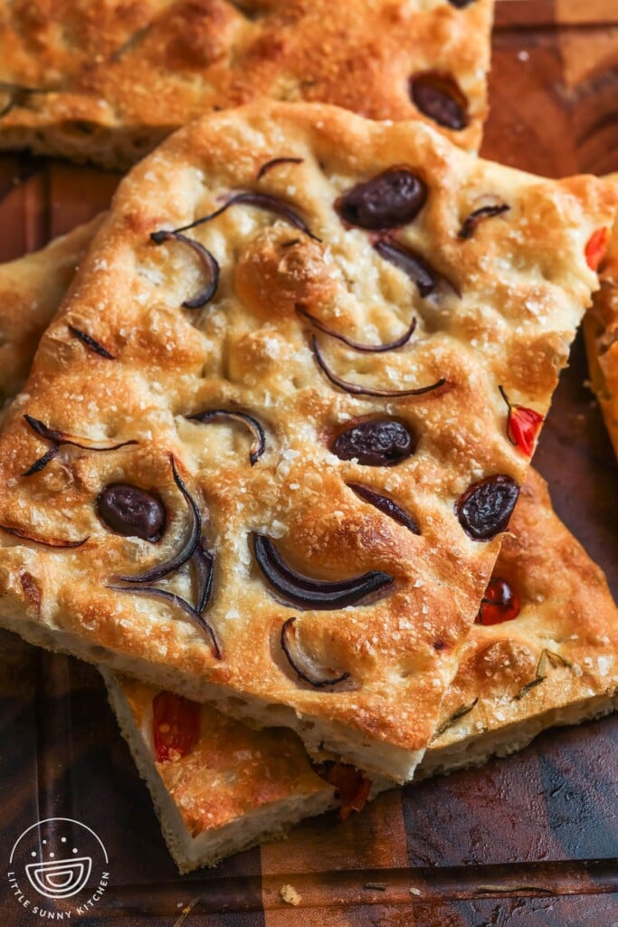 squares of thin sheet pan focaccia stacked on a wood board, topped with olives, onion, and flaky salt.