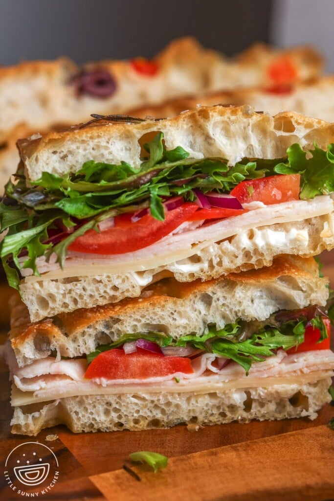 Two stacked focaccia sandwiches on a wooden board filled with sliced turkey, cheese, tomato, red onion, and greens