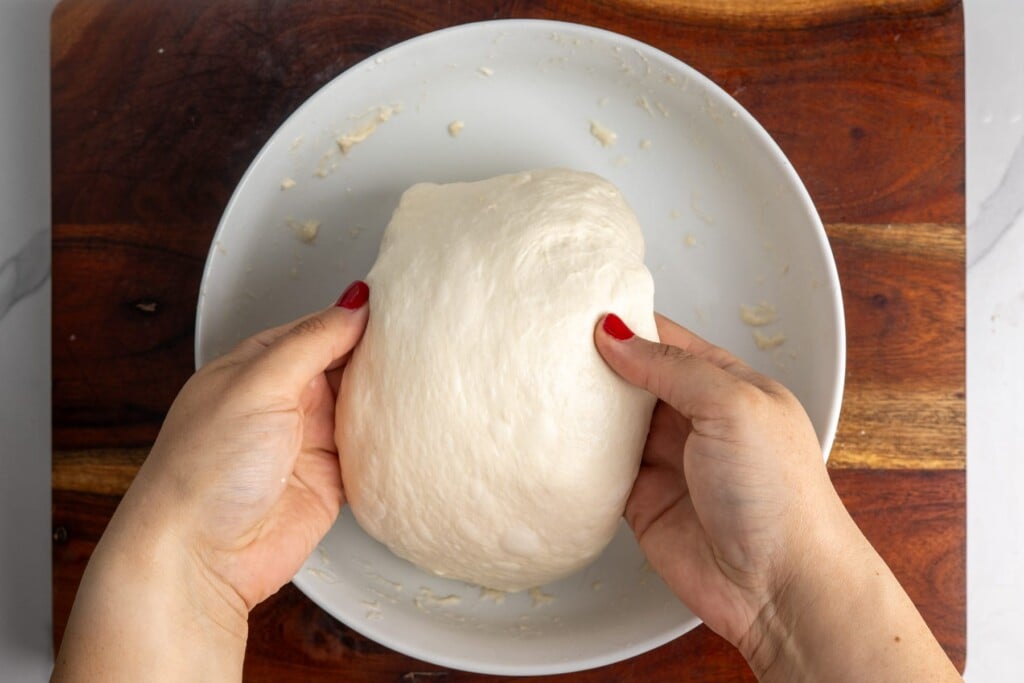 Two hands stretching one side of wet focaccia dough up and over the center of the bowl.