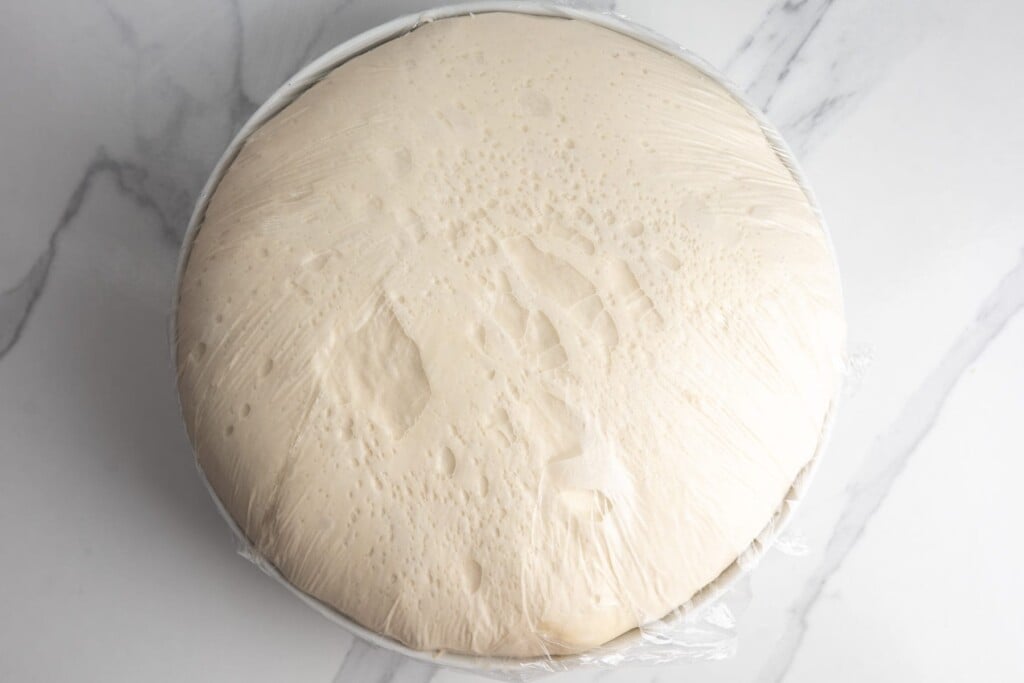 focaccia dough risen to the top of a bowl under plastic wrap after an overnight rise in the refrigerator. Showing bubbles under the surface.