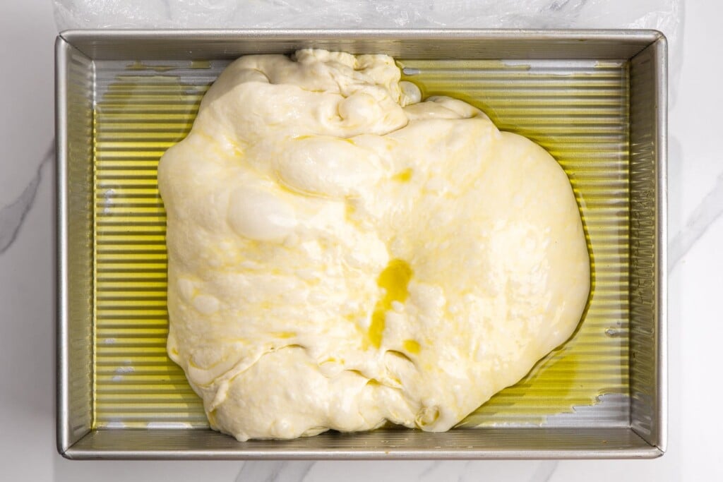 cold focaccia dough stretched in a metal pan coated with olive oil.