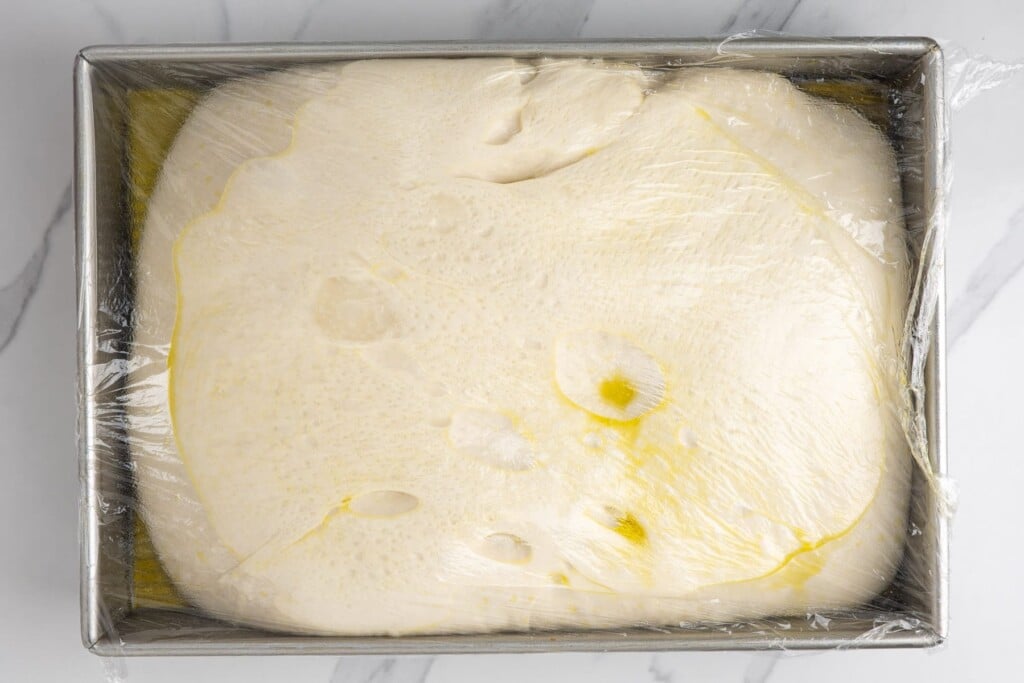 Proofed focaccia dough in a metal pan, puffy with large bubbles visible under the surface of the dough.