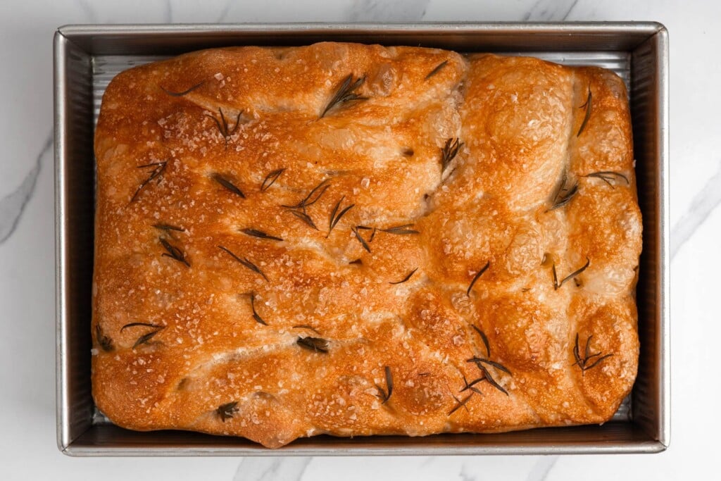 baked focaccia in a metal pan with deep golden top, with rosemary and sea salt on top.