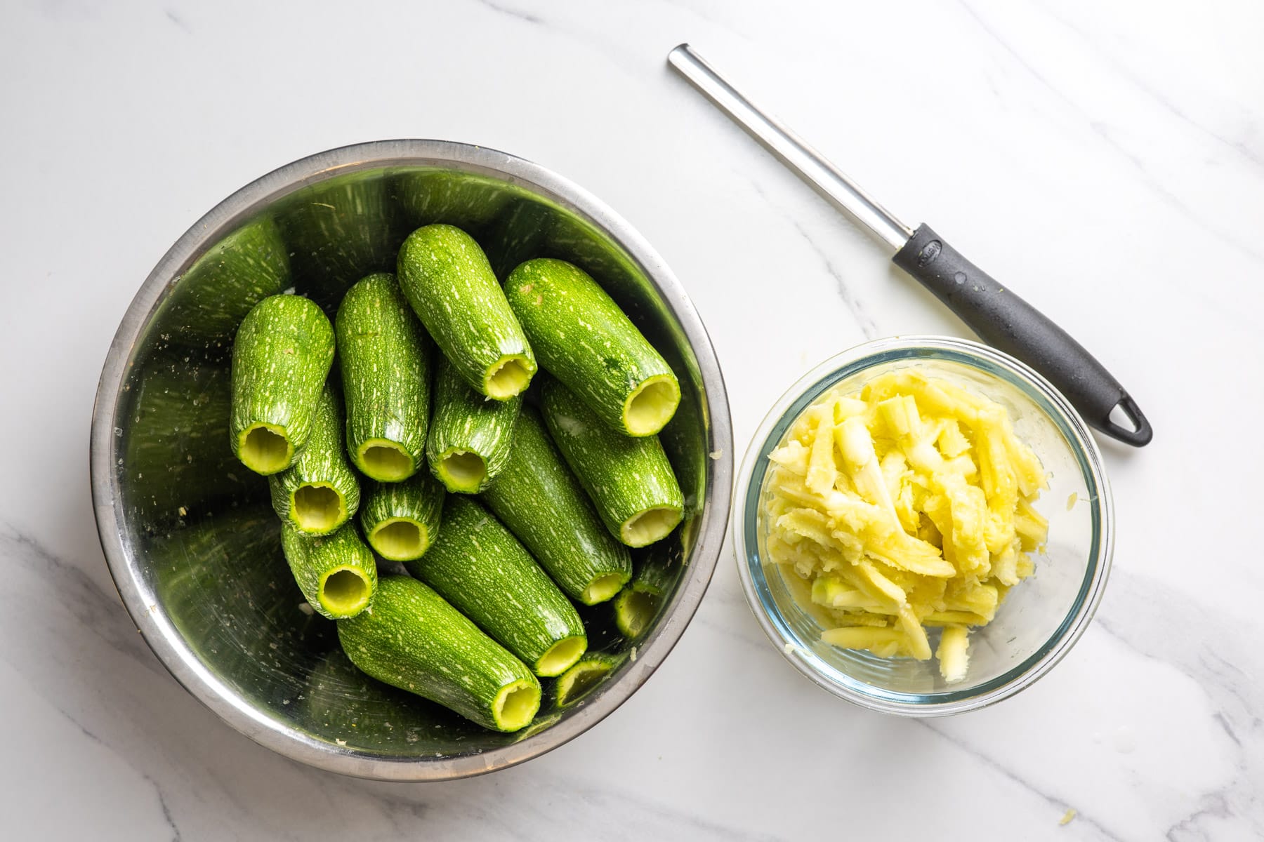 Hollowed out zucchini in a bowl next to a zucchini corer and the scooped out cores.