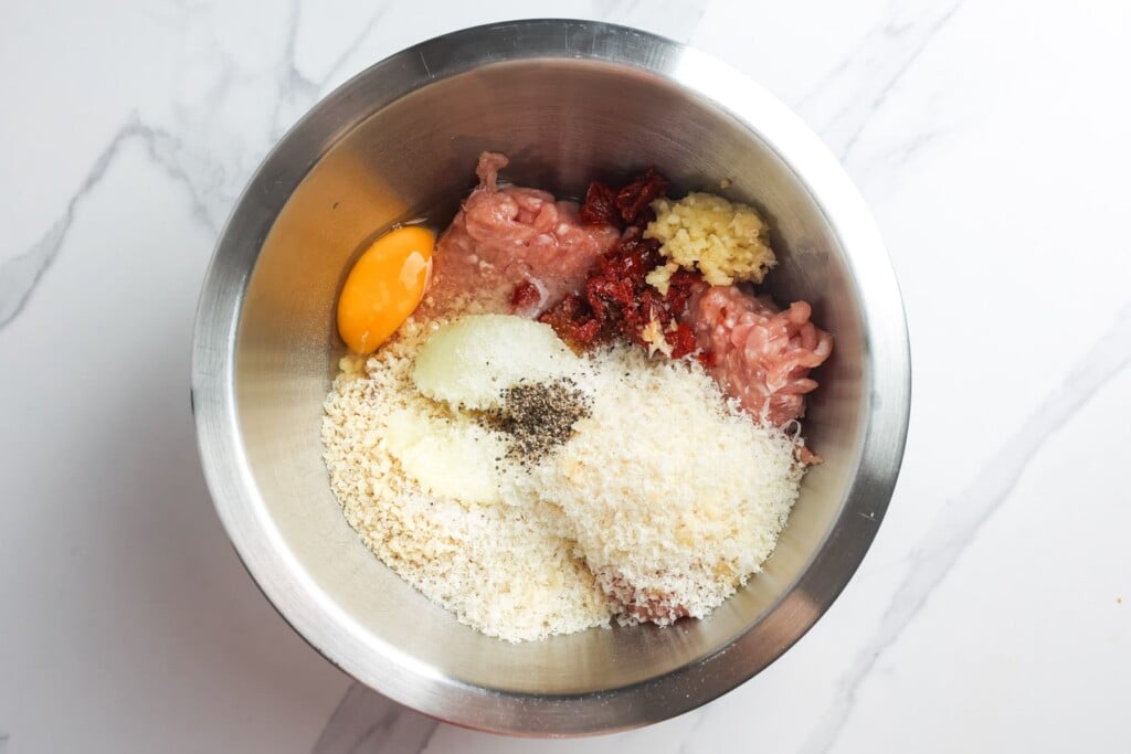 ground chicken in a metal mixing bowl with sun-dried tomatoes, grated onion, parmesan, breadcrumbs, egg, garlic, and seasonings for marry me chicken meatballs