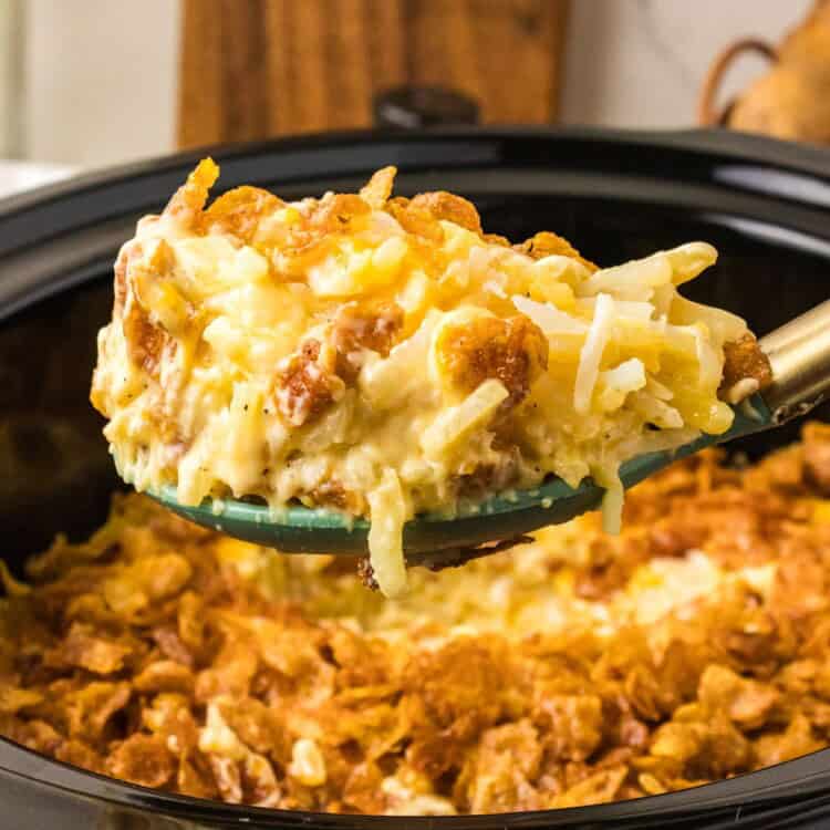 spoonful of crockpot cheesy potatoes lifted from a slow cooker, with crispy cornflake topping