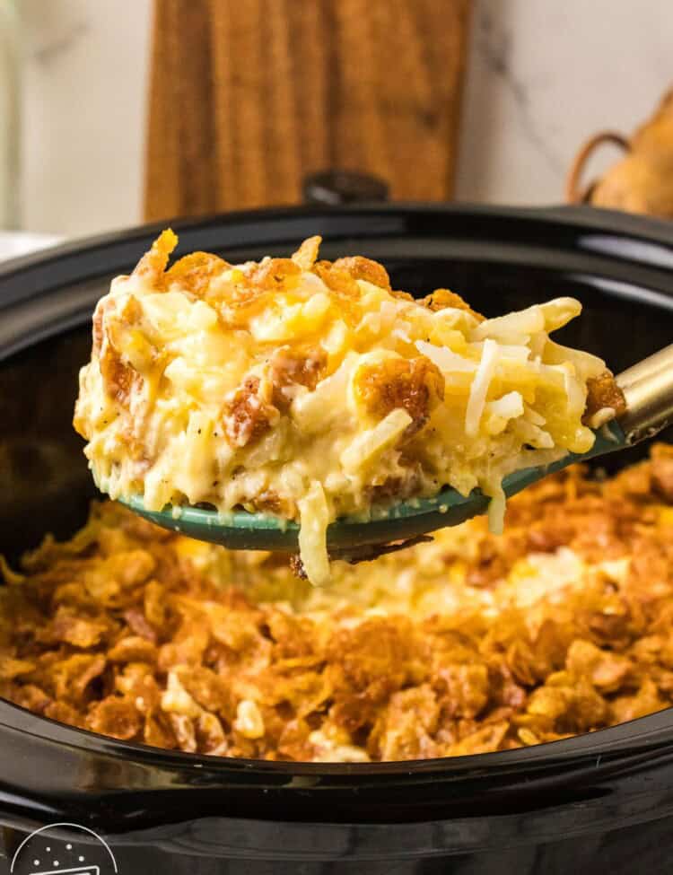 spoonful of crockpot cheesy potatoes lifted from a slow cooker, with crispy cornflake topping