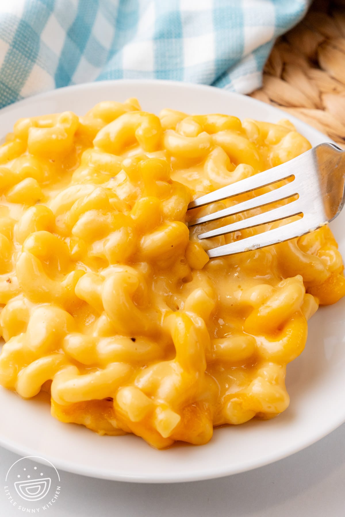 a serving of crockpot mac and cheese on a white plate with a fork, next to a blue and white checked napkin.