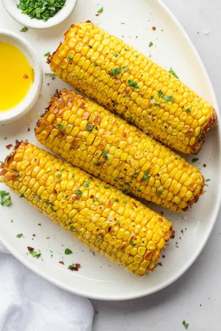 Three air fried corn on the cob on a white platter with melted butter and seasoned with chopped parsley and sea salt flakes.