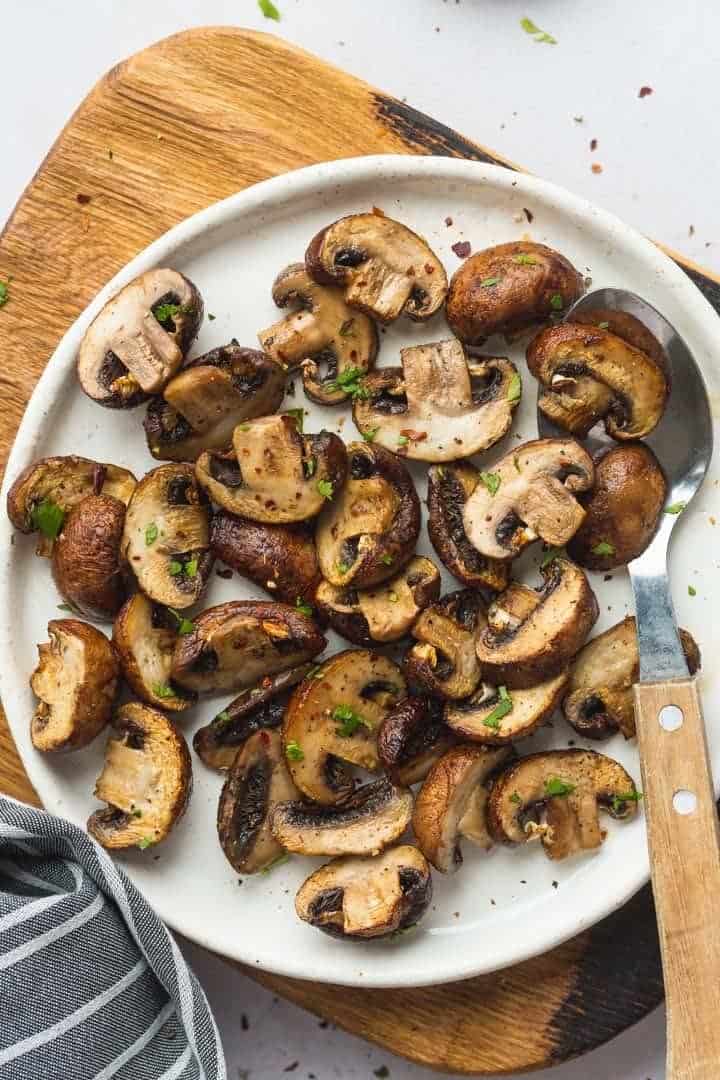 A white plate with air fryer mushrooms, on a wooden board, and a tea towel on the side.