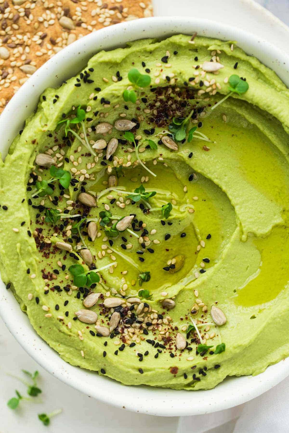 Avocado hummus in a white bowl, garnished with seeds, sumac, and microgreens.