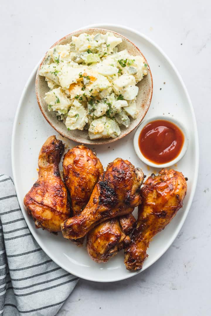 A platter of BBQ Chicken Drumsticks with potato salad, and ketchup - a top view shot of the final product.