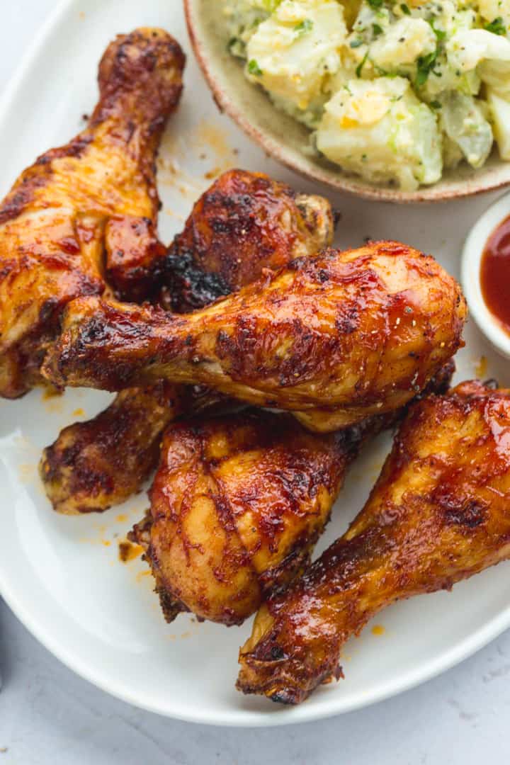 BBQ chicken drumsticks stacked on a white plate with potato salad side dish and red dip.