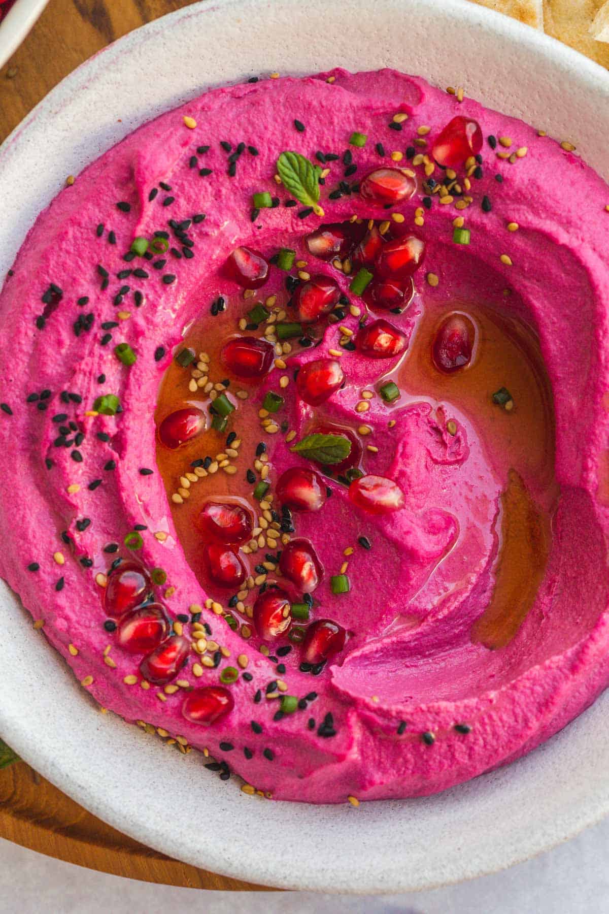 A close up shot of creamy beet hummus garnished with pomegranate seeds, sesame seeds, nigella seeds, mint leaves, chives, and olive oil