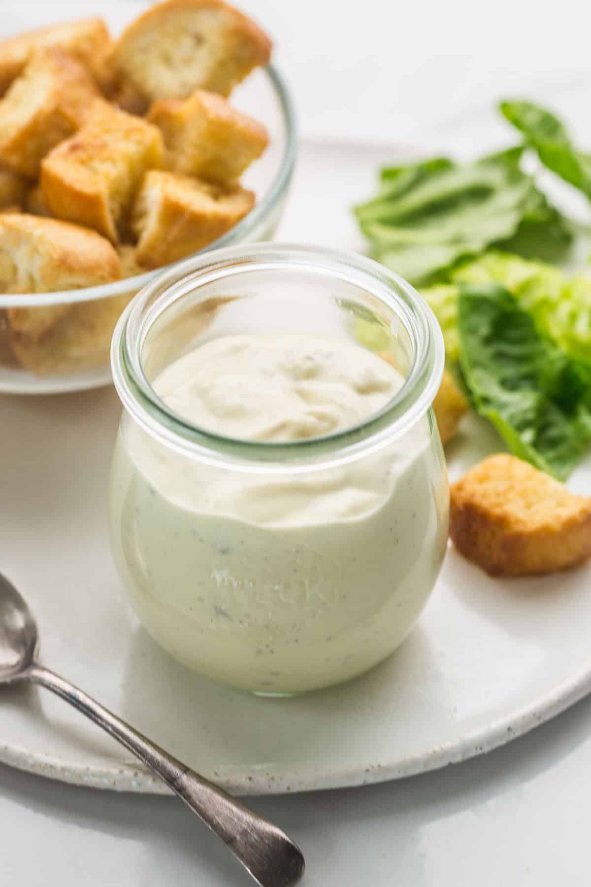 A small Weck jar with Caesar dressing on a ceramic white tray, and a spoon