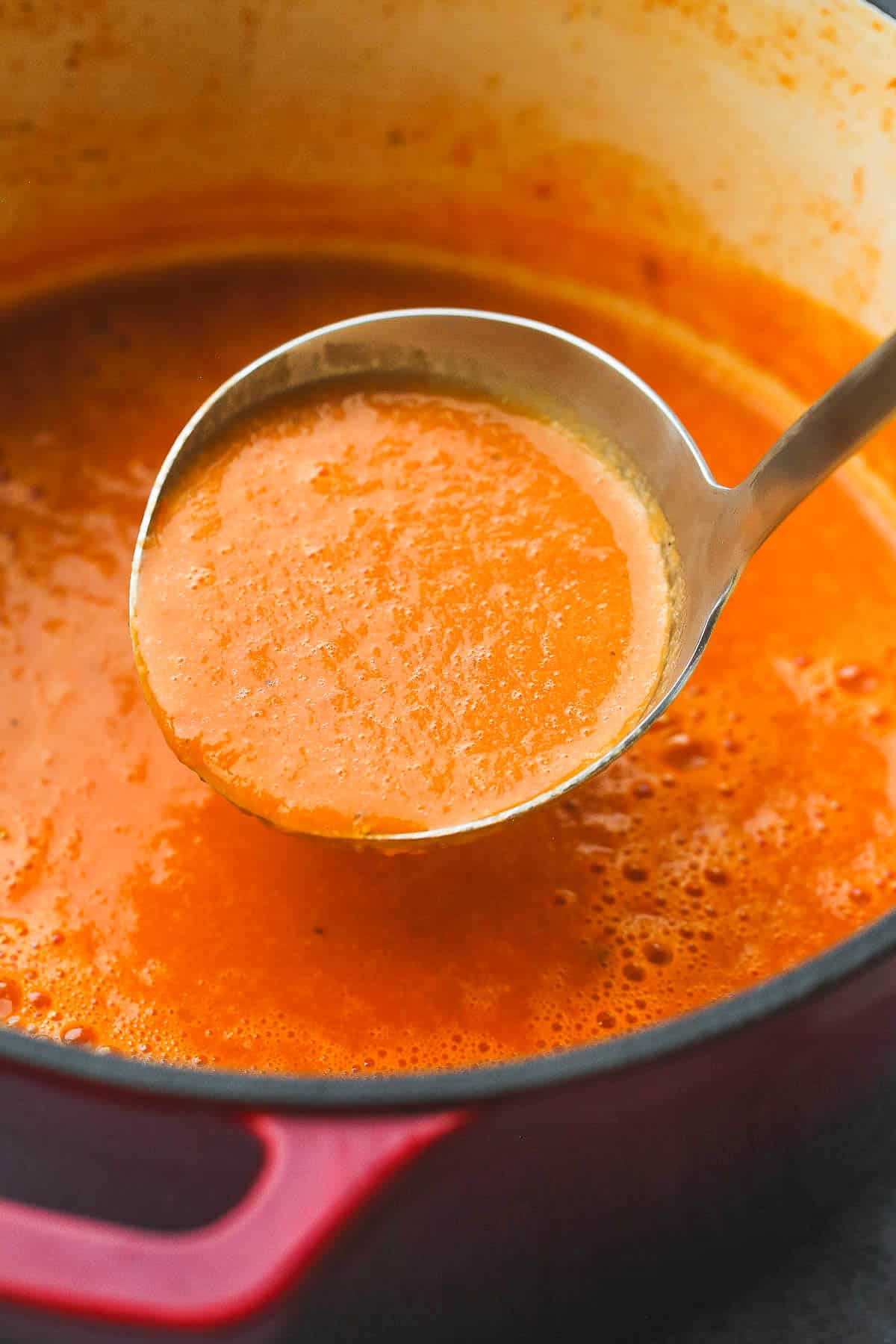 A metallic ladle with tomato soup and a red pot