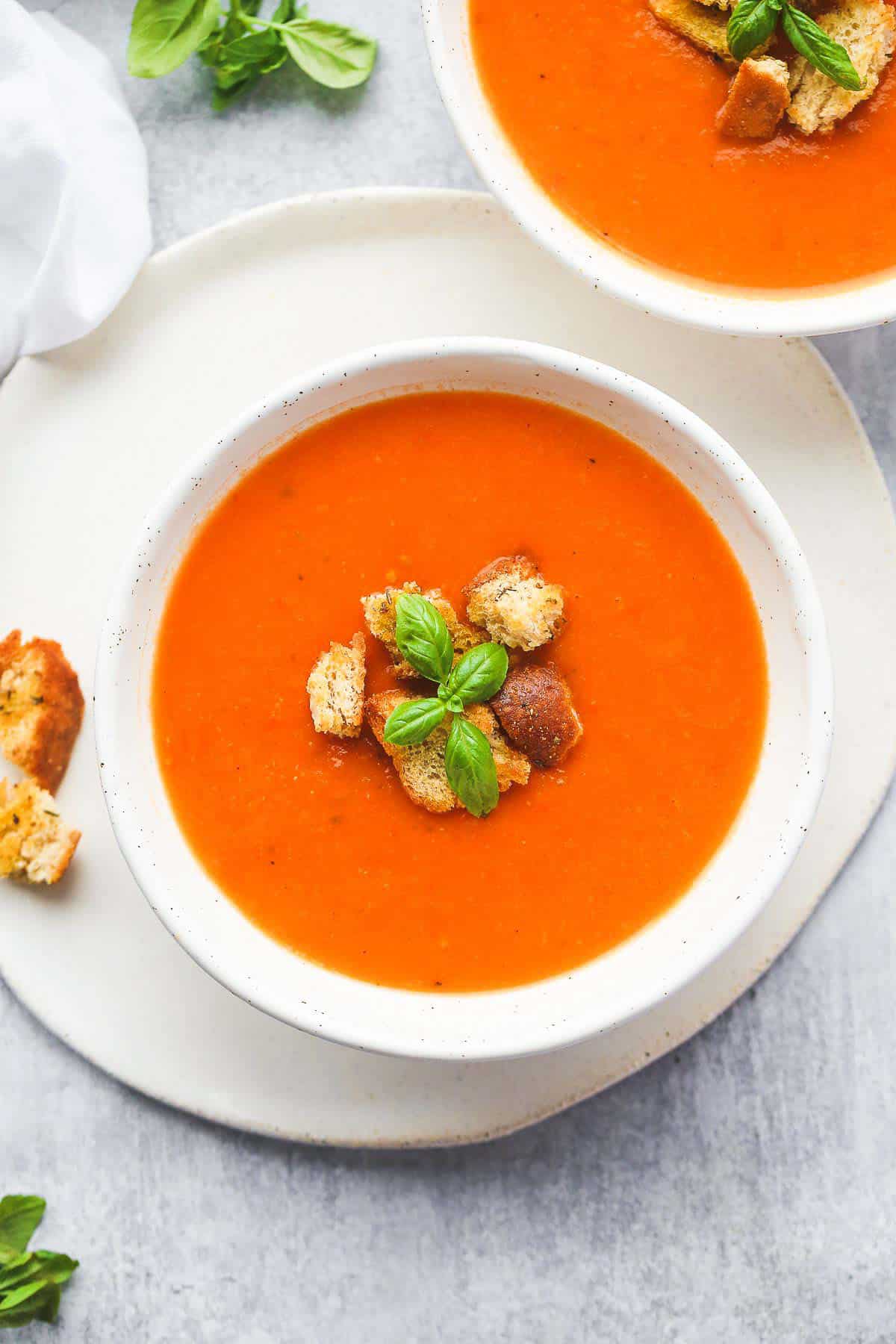 2 white bowls with tomato soup topped with croutons and basil leaves
