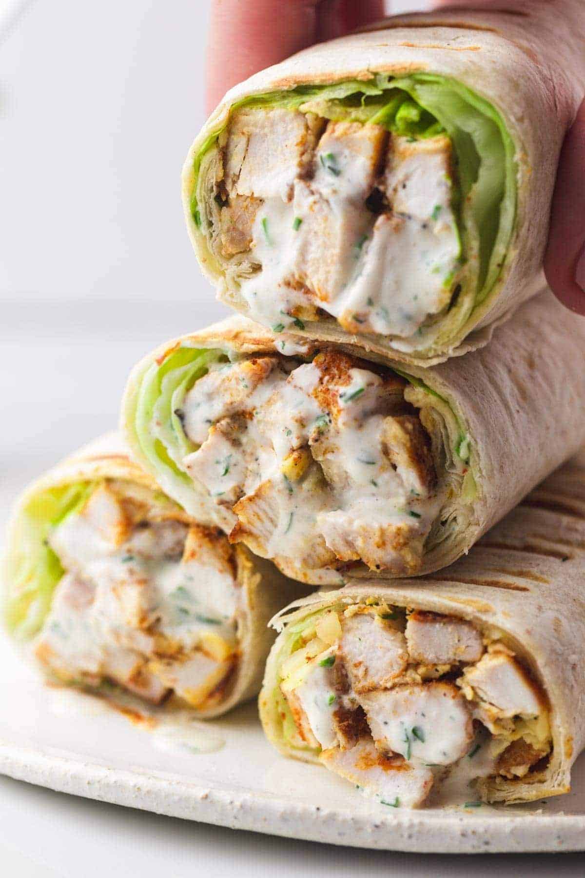 4 grilled chicken wraps stacked on each other, and a hand picking up the top wrap