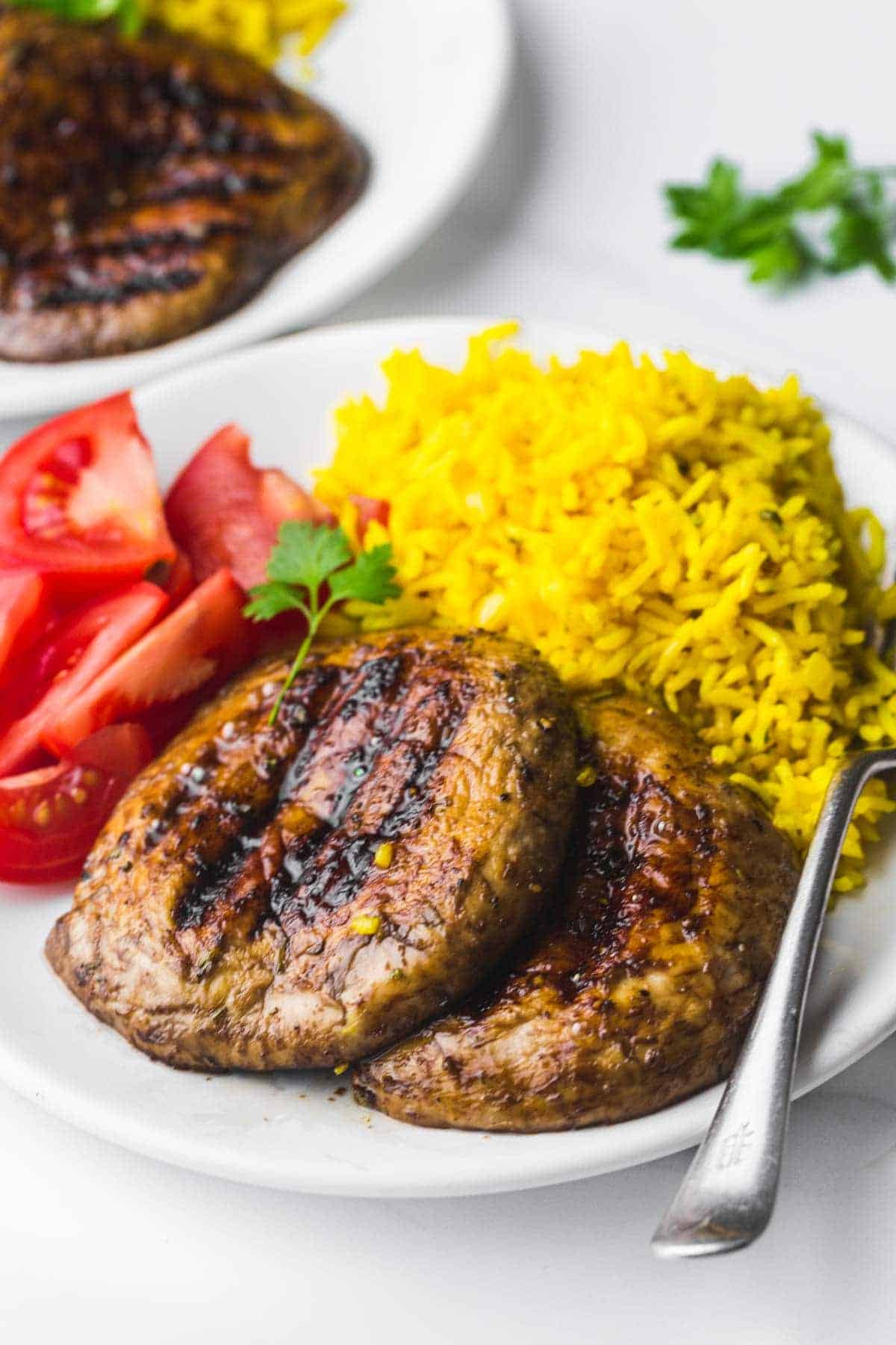2 grilled portobello mushrooms served on a plate with turmeric rice and sliced fresh tomatoes