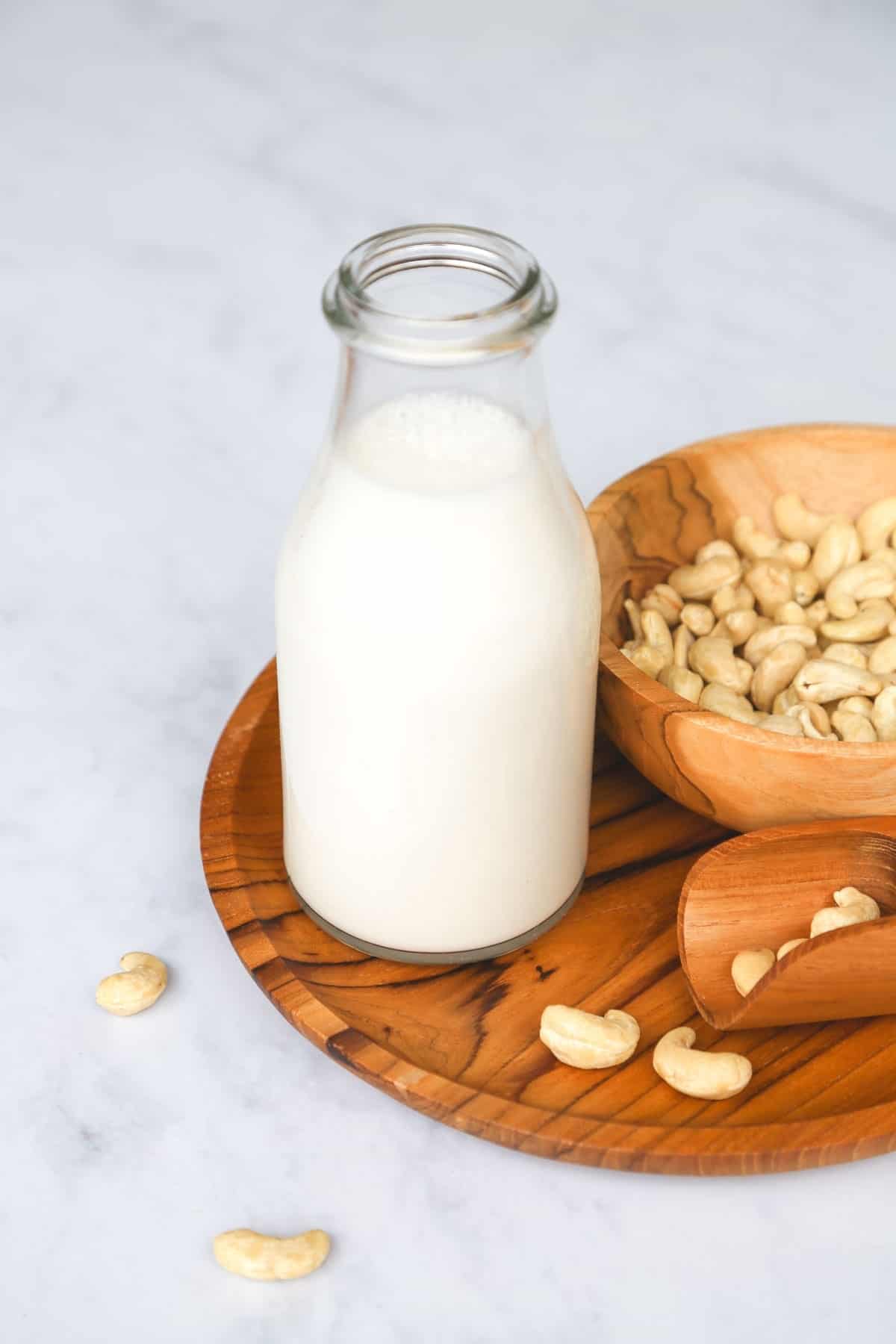 Homemade cashew milk in a small glass bottle, on a wooden tray, with some cashew nuts scattered on the tray
