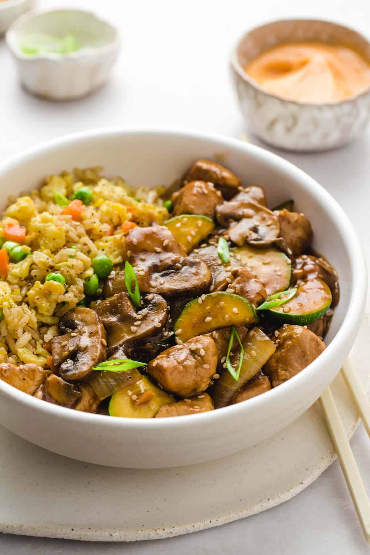 A white bowl with Hibachi chicken with zucchini, and fried rice. And little bowls with yum yum sauce and sliced green onion in the background