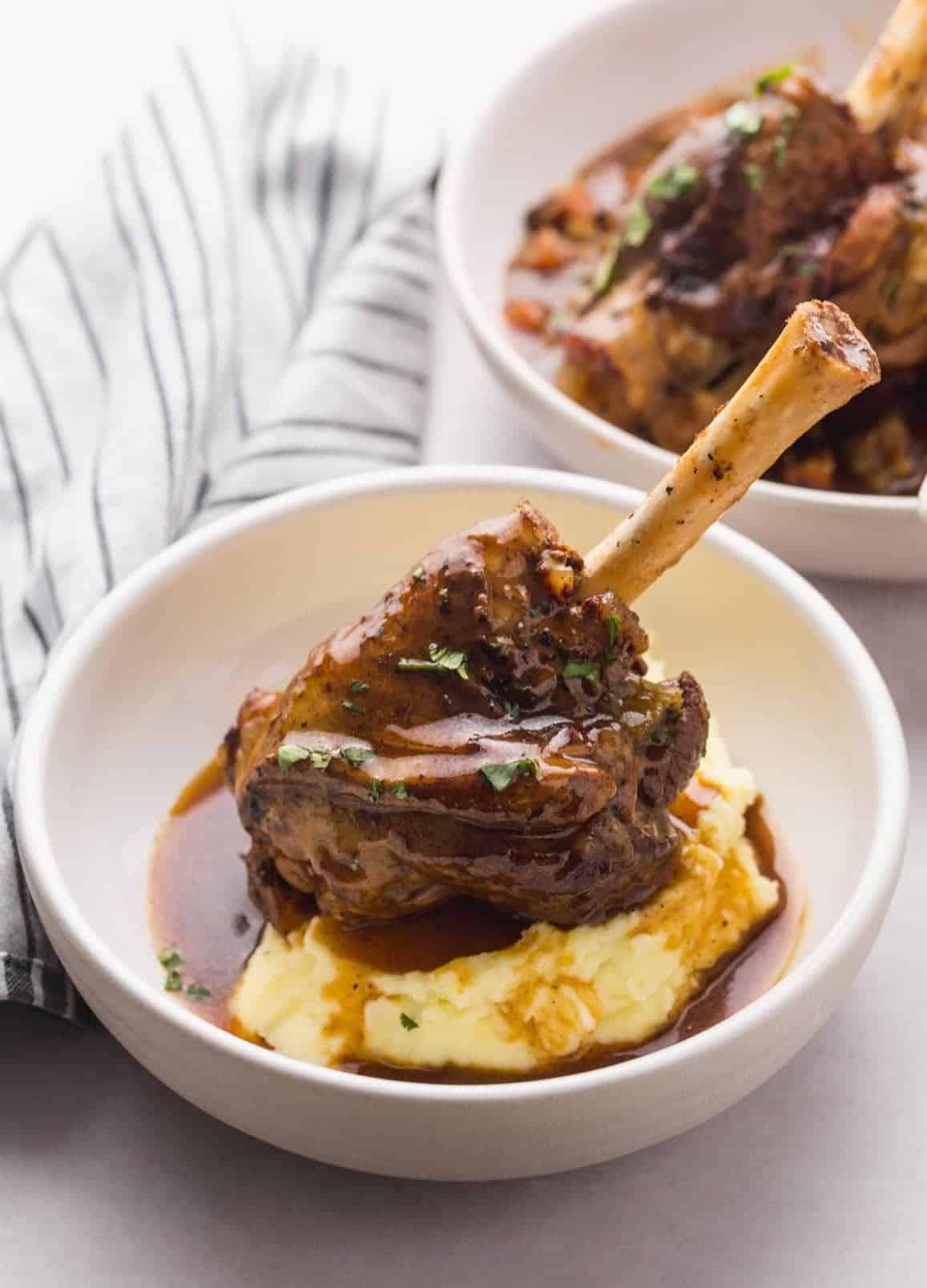 2 white plates/bowls with mashed potatoes and a braised lamb shank and gravy