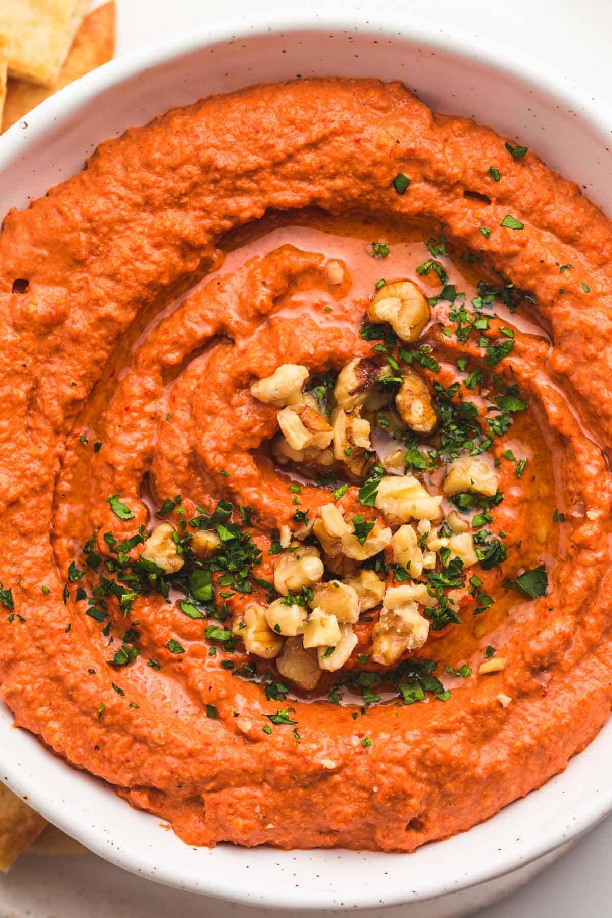 Close up of muhammara dip in a white bowl, with crushed walnuts and chopped parsley as garnish