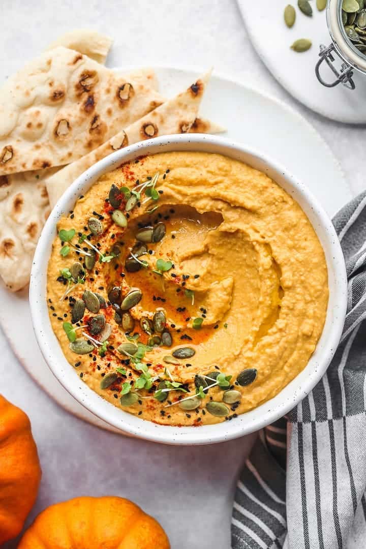 A flatly of a white bowl with pumpkin hummus, garnish with pumpkin seeds, watercress, nigella seeds and smoked paprika.