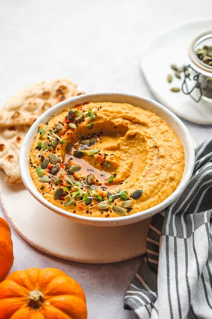 A white bowl with pumpkin hummus garnished with olive oil, pumpkin seeds, watercress, nigella seeds and smoked paprika.
