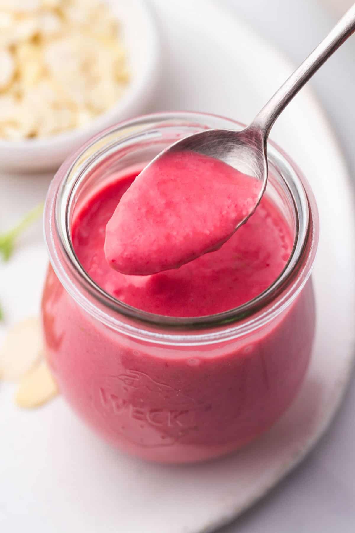A small Weck jar filled with Raspberry Vinaigrette, and a teaspoon filled with the vinaigrette to show the texture