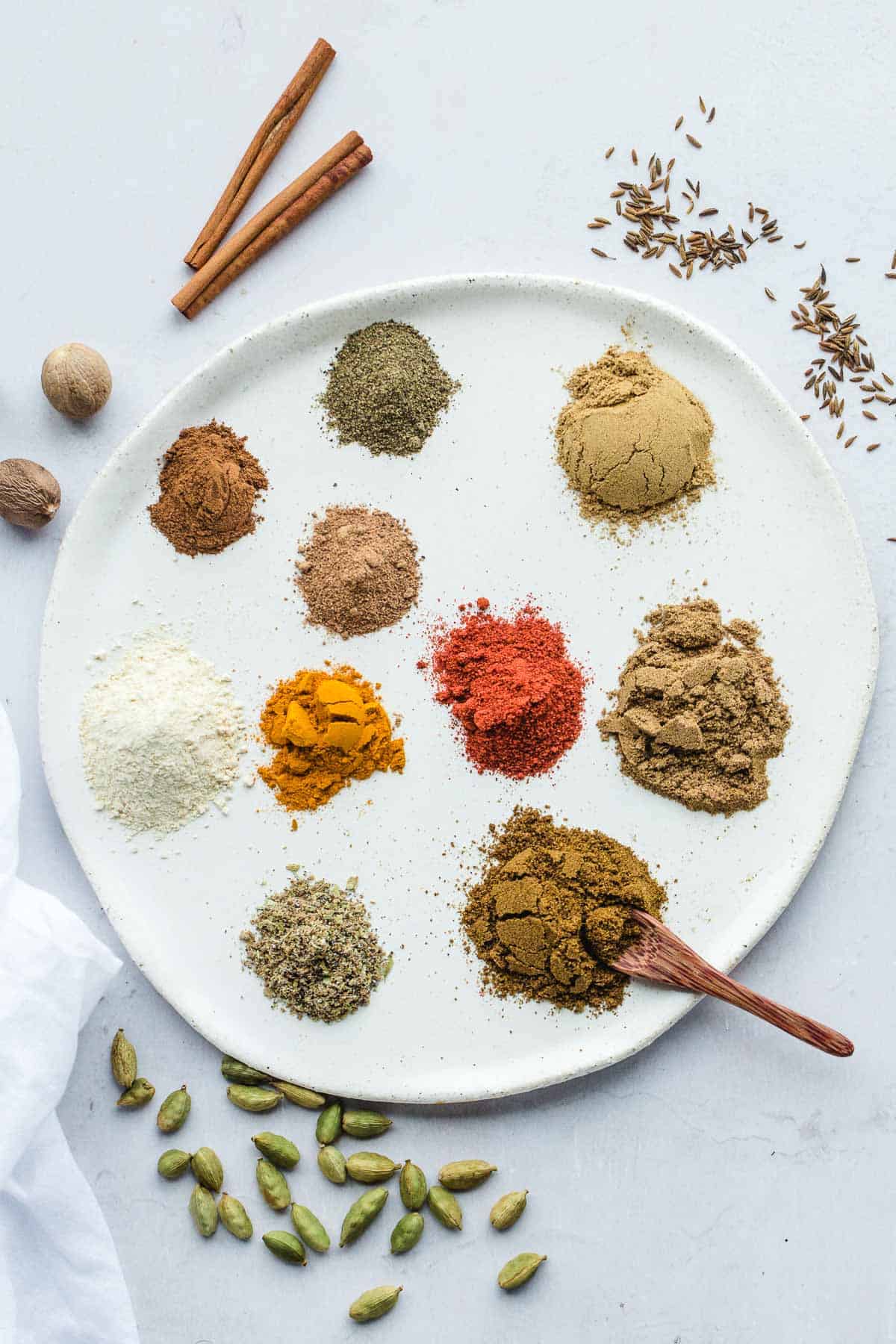 All the ground spices laid out of a round ceramic white tray, along with whole spices on the sides, and a small wooden spoon