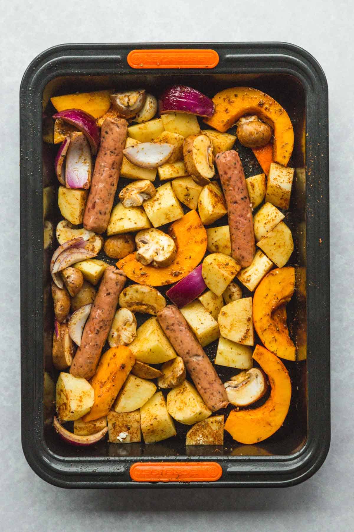 Chopped veggies and sausages in a roasting tin with seasonings and olive oil