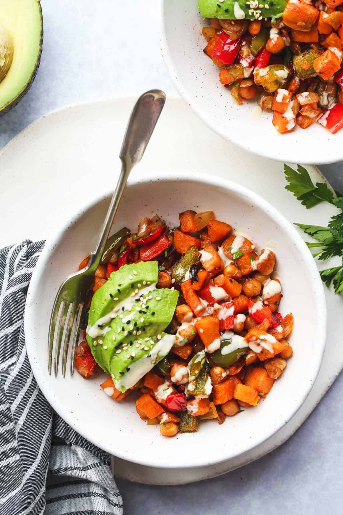 A white bowl eith sweet potato hash, fresh avocado slices, and tahini dressing drizzle. With a fork on the side, and a kitchen tea towel in the background.