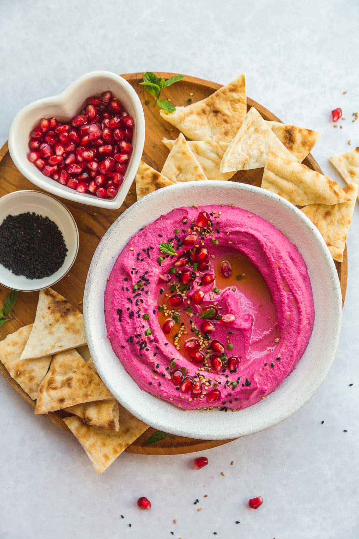 Creamy Beet Hummus in a white bowl, with small bowls with pomegranate seeds and nigella seeds
